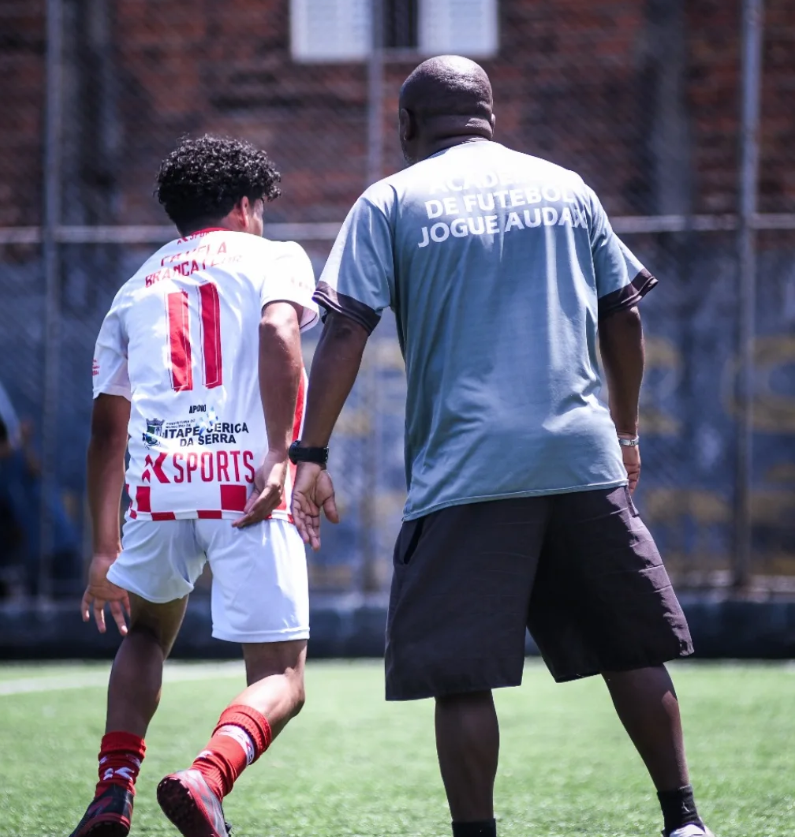 Um adulto e um jovem atleta caminham juntos em um campo de futebol, vistos de costas.